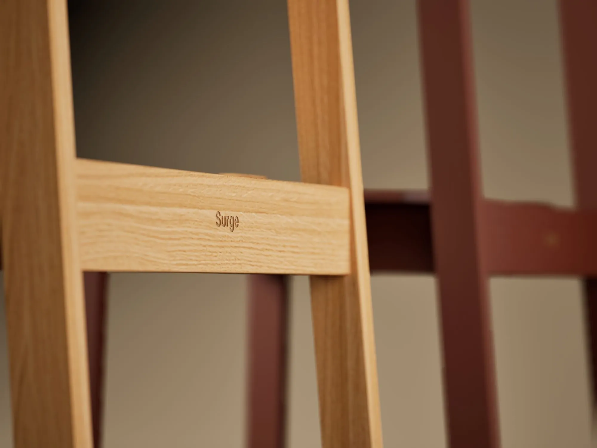 Close-up of an oak chair frame with the word Surge engraved, a red chair out of focus behind